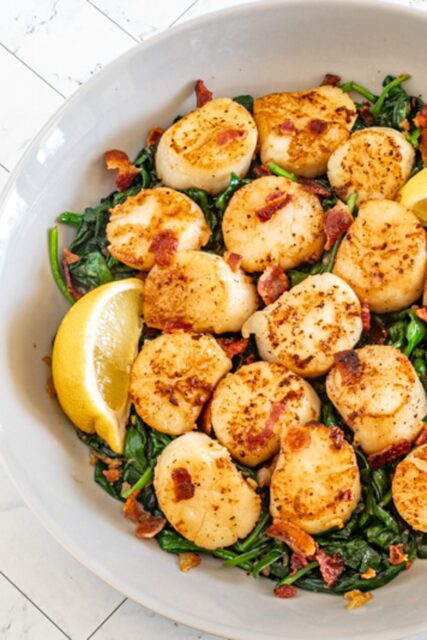 scallops on a bed of spinach with lemon wedges and bits of bacon