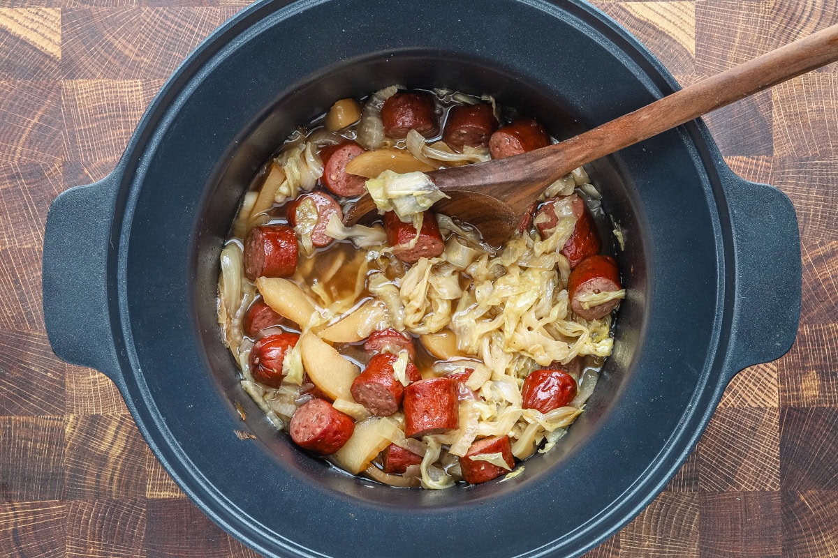 A slow cooker with a cooked mixture of cabbage, apples, and kielbasa smoked sausage, with a wooden spoon for serving.