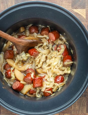 Slow cooker cooked kielbasa with apples, cabbage, onions, and tangy sauce, shown in the crock pot with a wooden spoon.