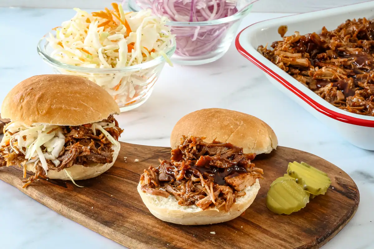 2 small pulled pork sandwiches wit a dish of pulled pork , onion, and coleslaw in the background.
