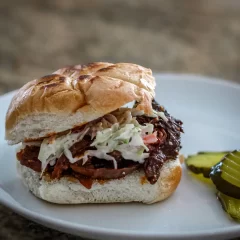 Slow-cooked shredded beef sandwich with coleslaw on a bun, garnished with pickles.