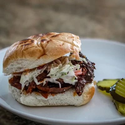 Slow-cooked shredded beef sandwich with coleslaw on a bun, garnished with pickles.