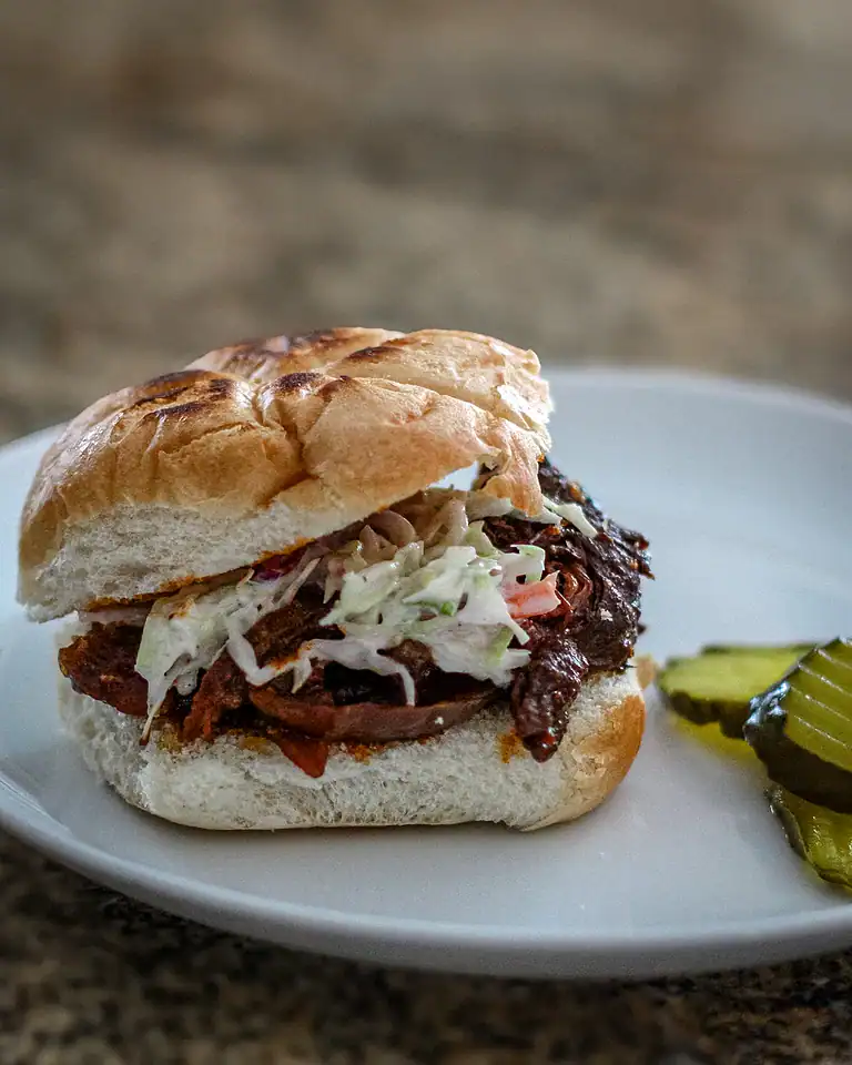 Slow-cooked shredded beef sandwich with coleslaw on a bun, garnished with pickles.