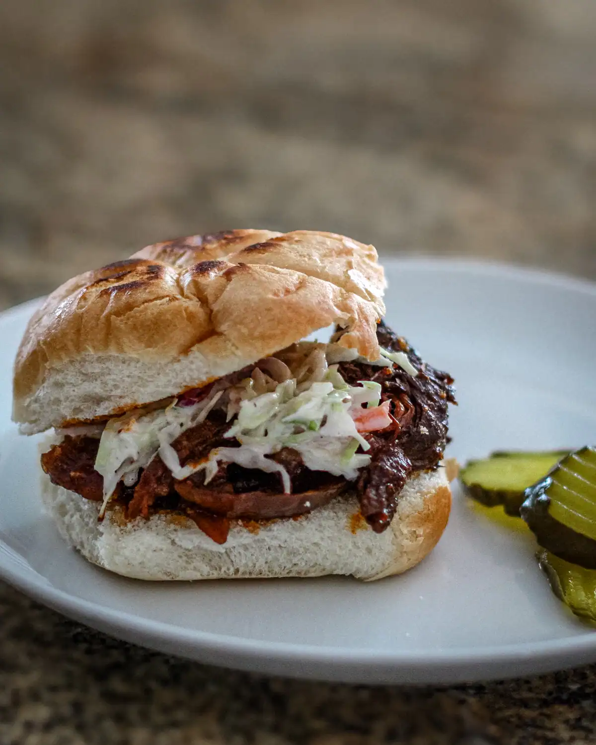 Slow-cooked shredded beef sandwich with coleslaw on a bun, garnished with pickles.