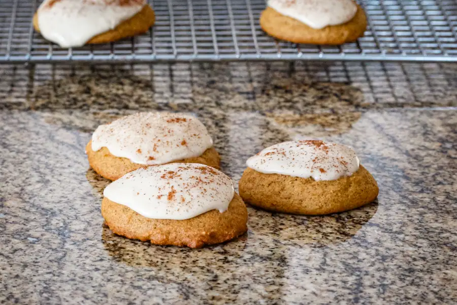 Iced pumpkin cookies on a kitchen counter with more on a cooling rack in the background.