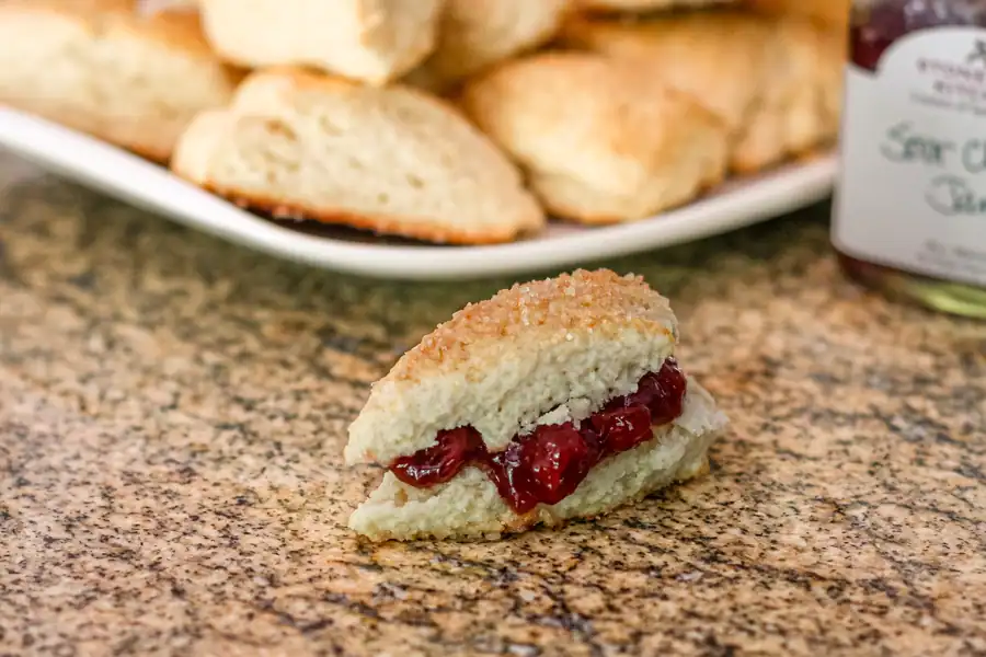 A vanilla scone with cherry preserves and a serving plate with more scones in the background.

